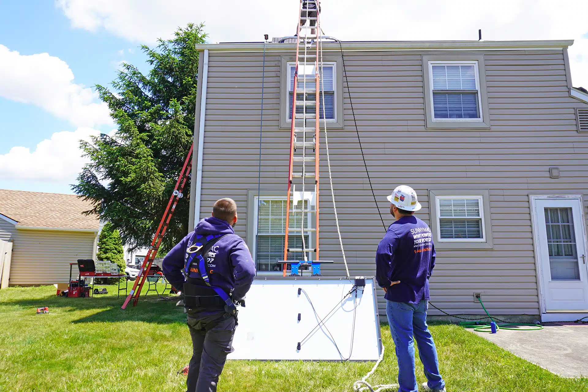 Two SunnyMac employees stand outside house with ladder while another employee works on the roof